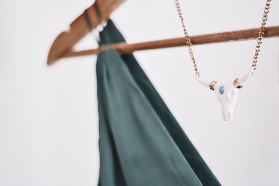 A bull skull necklace hangs on a hanger with a green silk dress on a white background.