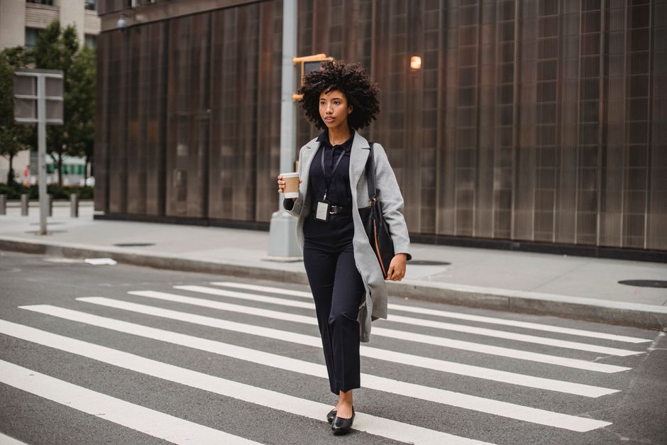 Young ethnic female employee with badge and flying hair crossing city road with coffee to go while looking forward