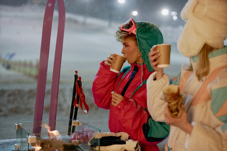 Two women in winter attire enjoying hot beverages by the ski slopes at night.