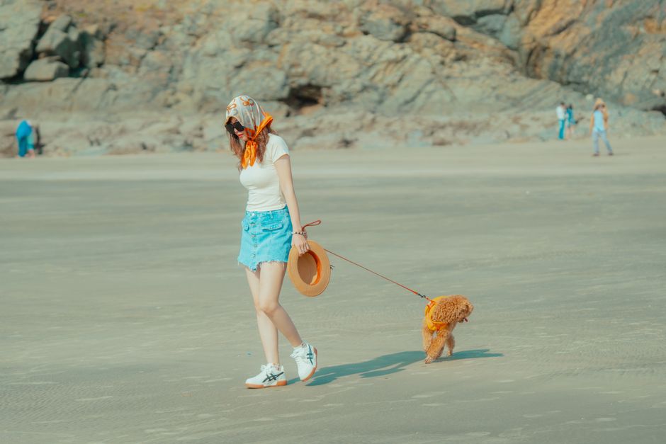 A woman in casual summer attire walks her dog on a scenic beach with rocky cliffs.