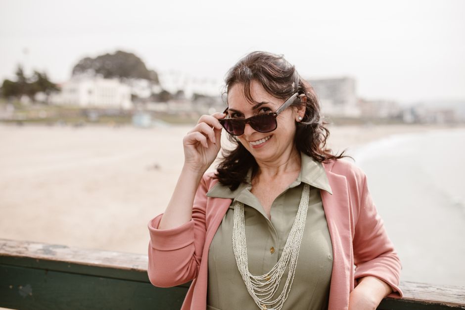 A stylish woman in a pink blazer smiles while adjusting her sunglasses at a sunny beach.