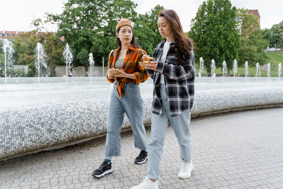 Two women in denim and plaid enjoy food while walking by a fountain in a park.