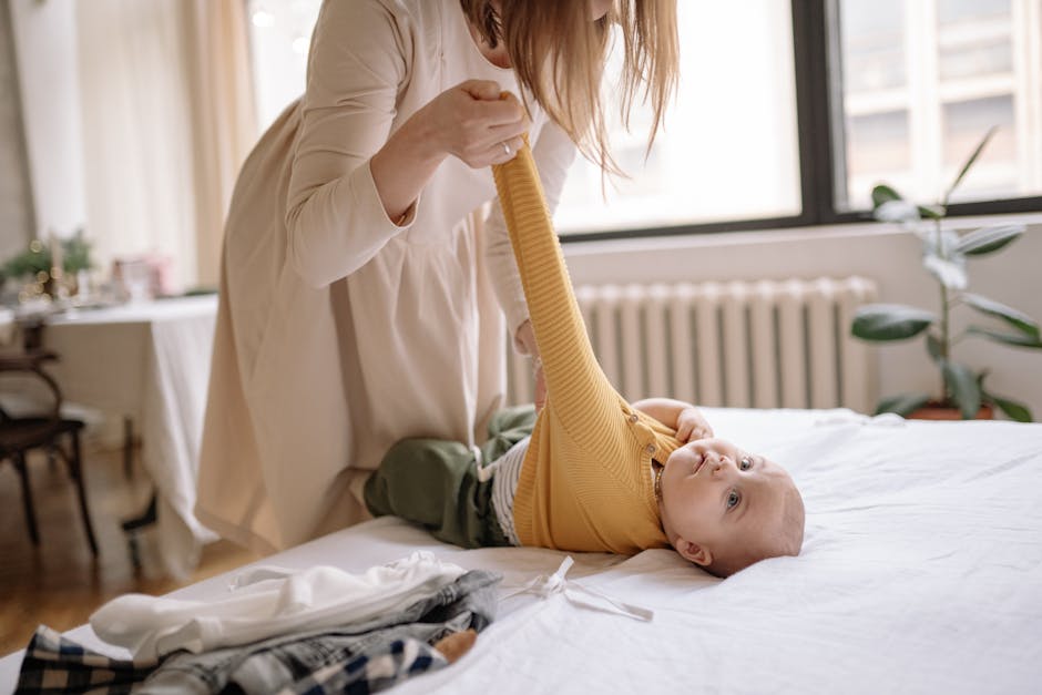 A mother helps her baby into a yellow sweater in a warm, sunlit bedroom.