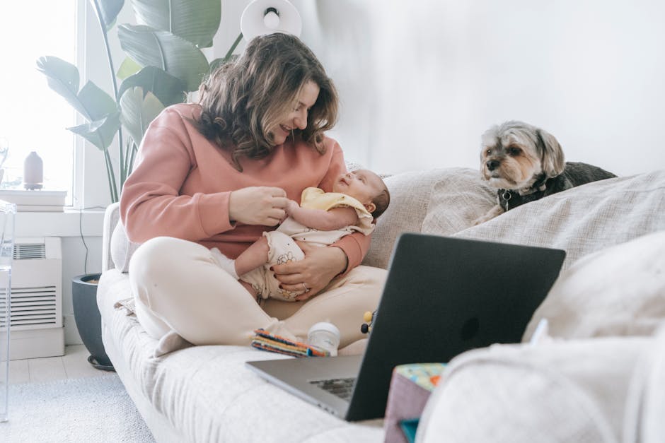 Full body of content mother with cute newborn baby in arms sitting on comfortable sofa with netbook near dog at home