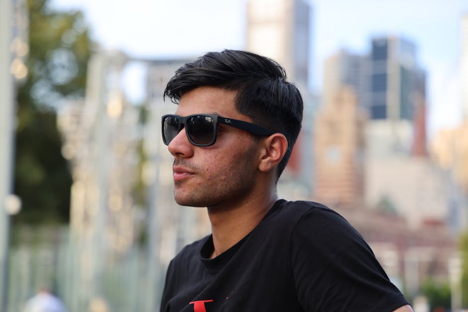Stylish young man wearing sunglasses, captured outdoors with a blurred cityscape background.