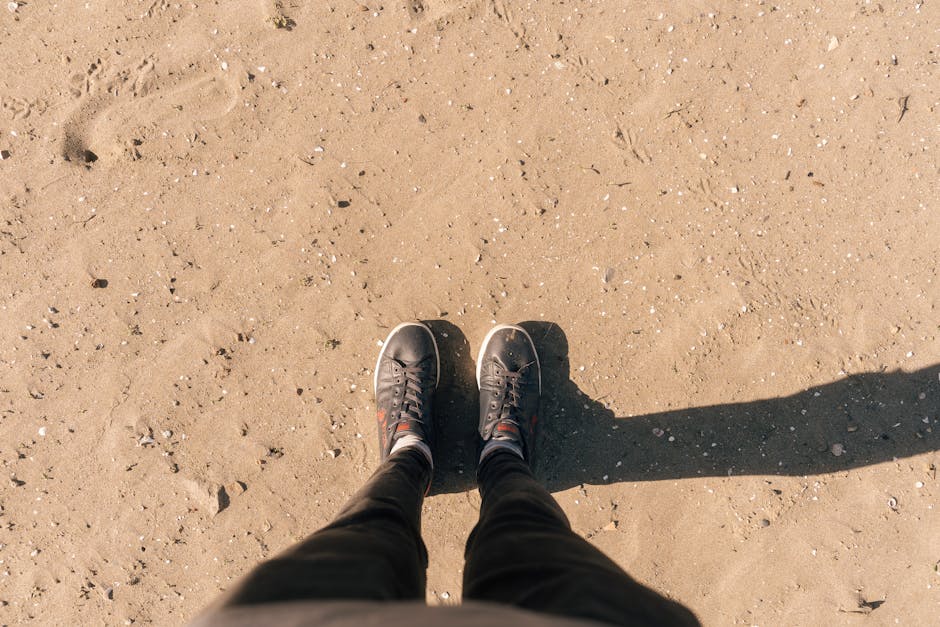 View of a person's shoes standing on a sandy beach, casting shadows.