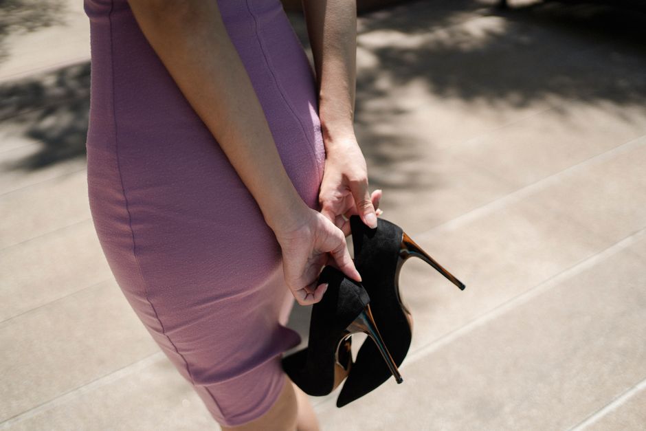 From above of crop faceless young lady in stylish dress holding high heeled shoes in hands while standing on street on sunny day