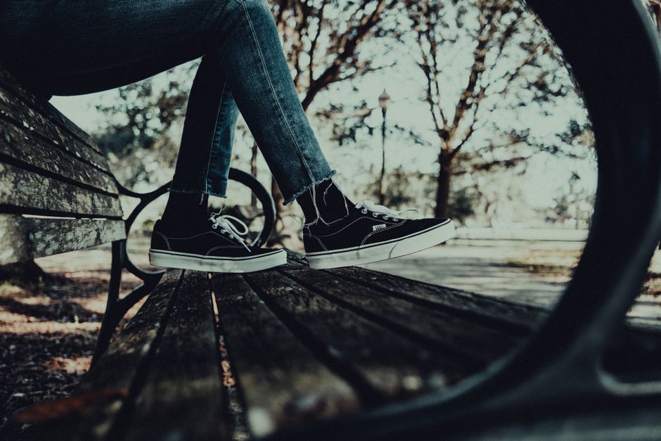 Close-up of black sneakers and denim jeans on a wooden bench in a park setting.