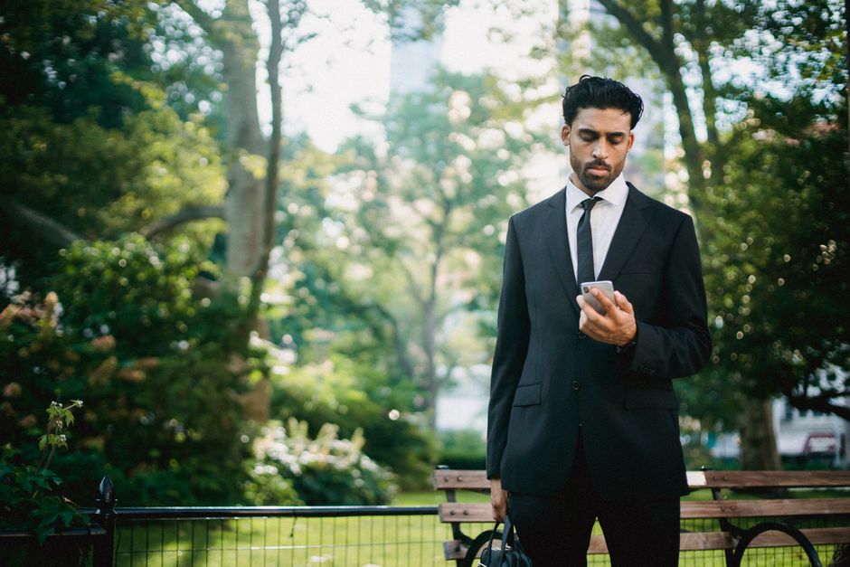 Confident businessman in a black suit checks his phone in a lush city park, blending professional elegance with a natural setting.