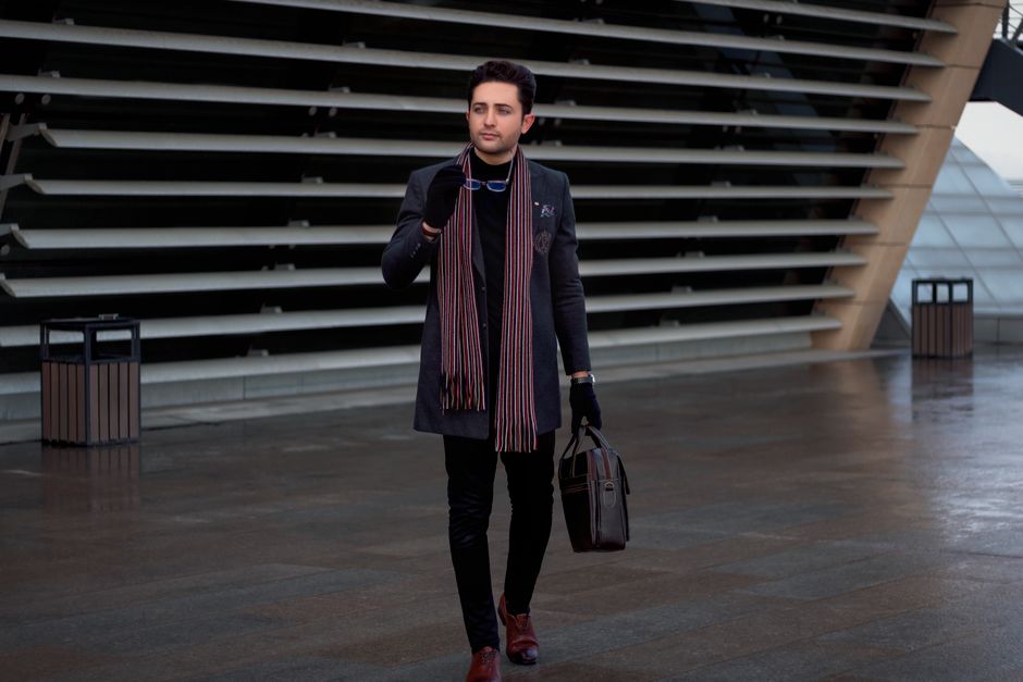 A fashionable man with a briefcase and scarf walking near modern architecture in Tehran.