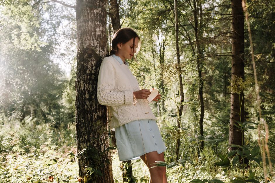 A woman in a knitted sweater leans against a tree reading a letter in a serene forest setting.