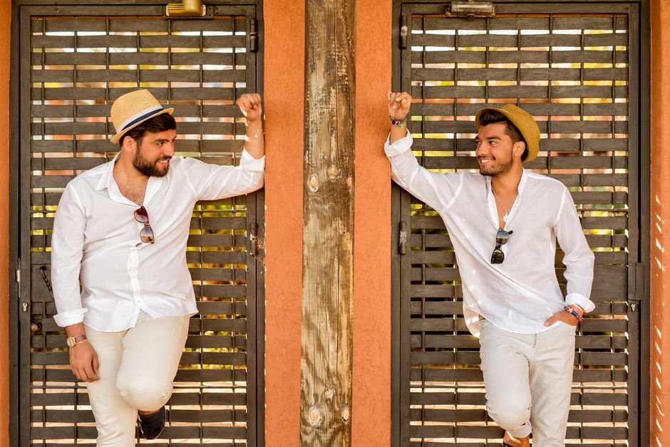 Two stylish men in straw hats and sunglasses lean on a rustic gate, sharing smiles.