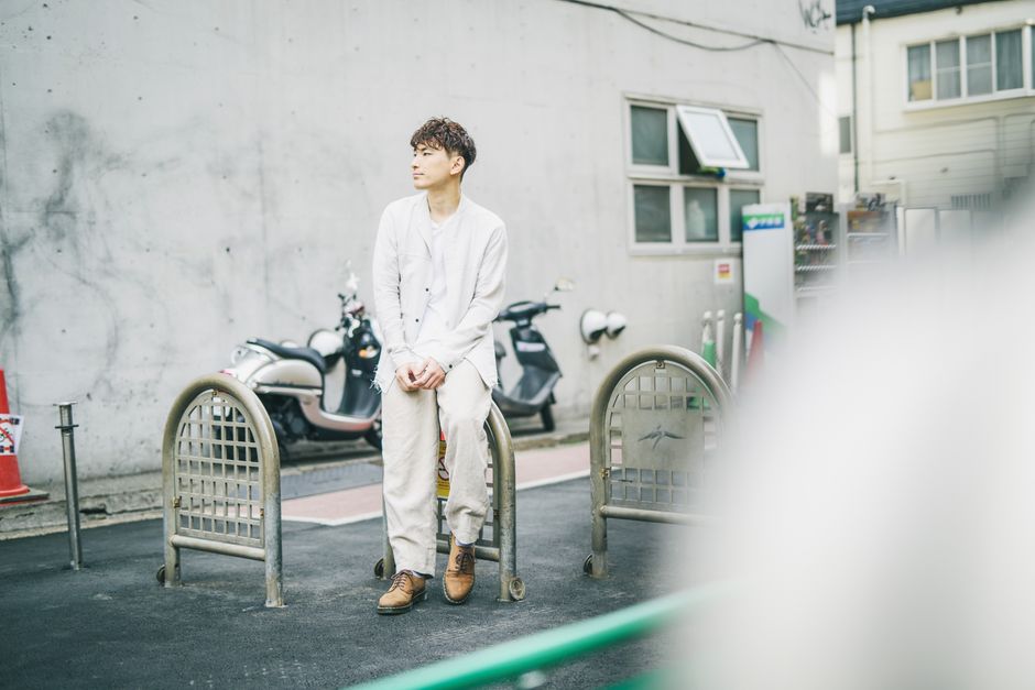 Asian man wearing white suit sitting on a sidewalk with motorbikes in the background.