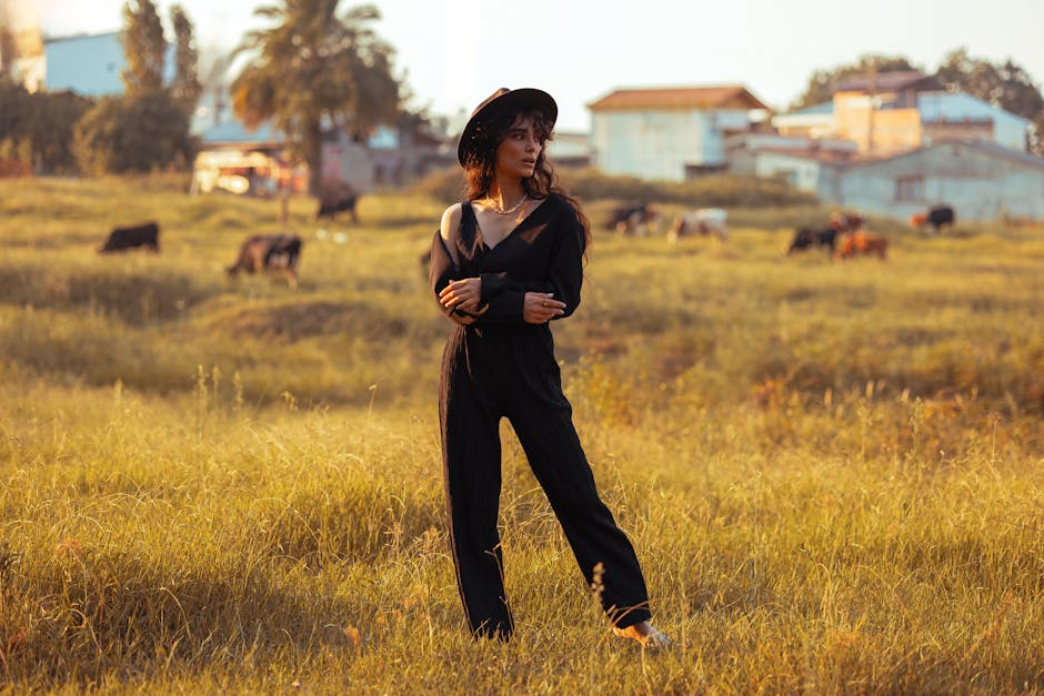 Stylish woman in black attire poses in sunlit field with cows in background, exuding rustic charm.