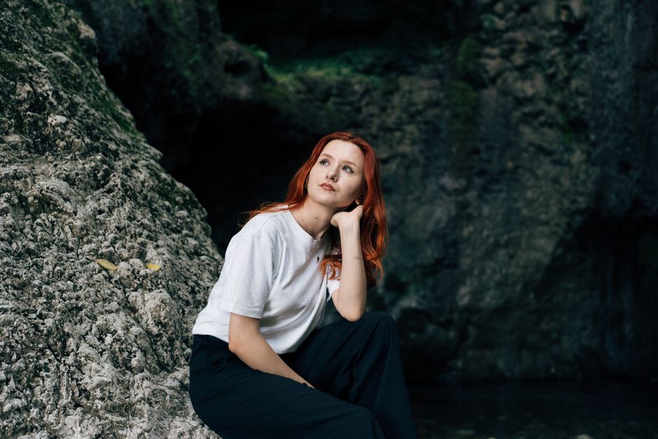 A thoughtful woman with red hair sits by rocks, capturing a serene and introspective moment.