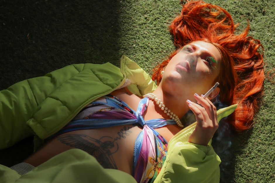 A stylish individual with red hair relaxes outdoors, smoking a cigarette on a sunny day.