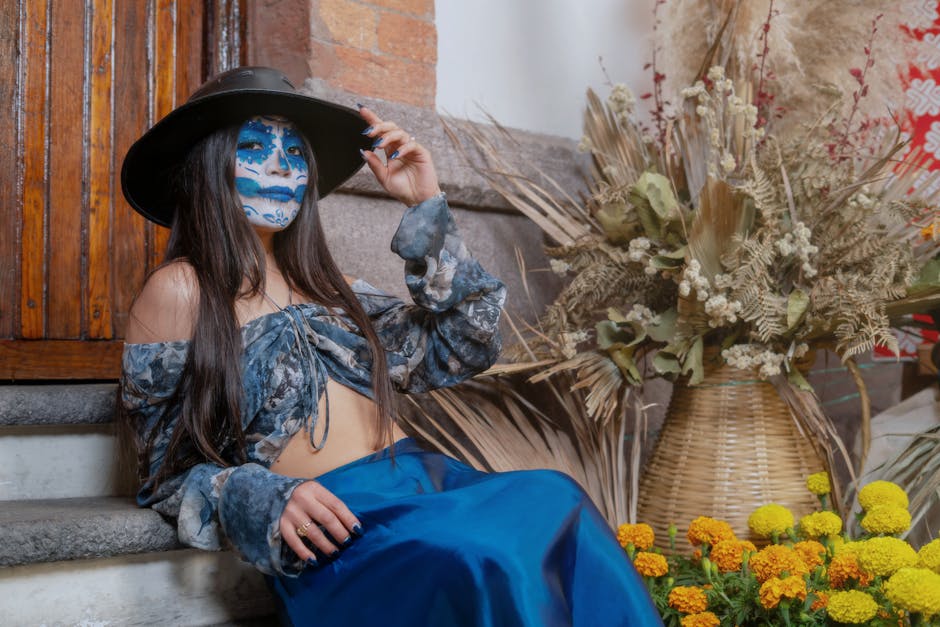 Woman with Day of the Dead makeup sitting beside vibrant flowers indoors.