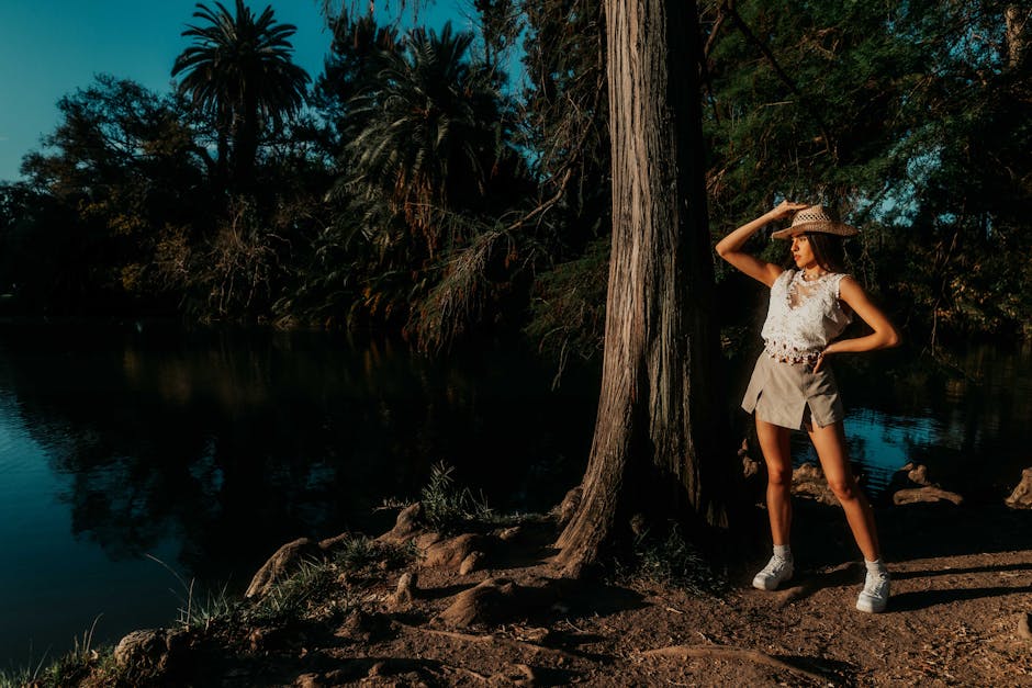 Woman in stylish attire posing by a lake in a sunny, natural setting.