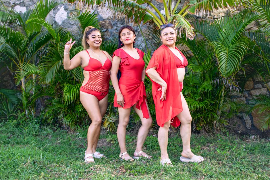 Three women in red swimwear standing outdoors with green foliage background, smiling confidently.