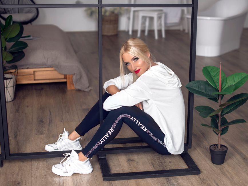 Blonde woman in white hoodie and leggings sitting indoors by a mirror.