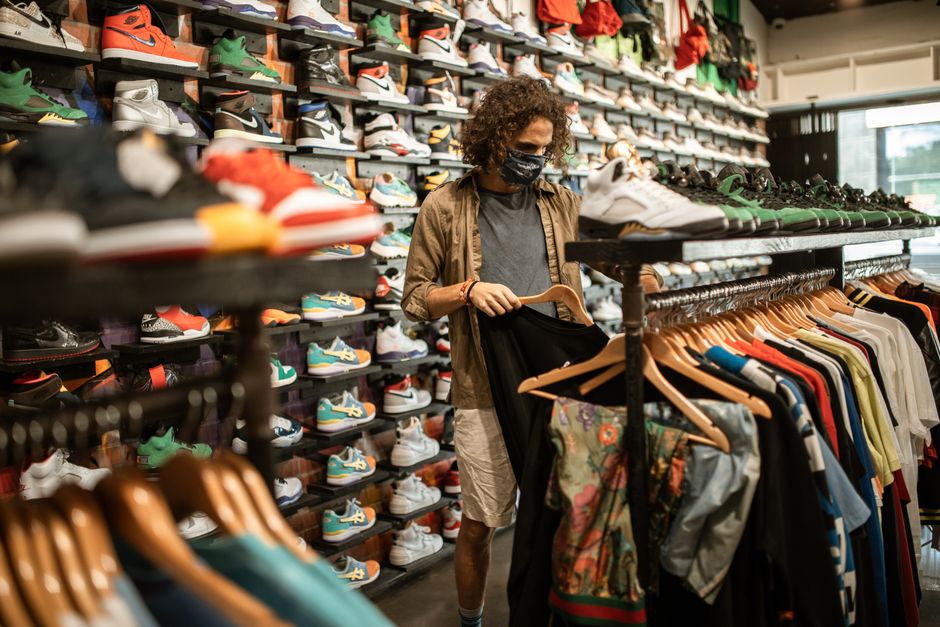 Young man in a sneaker and clothing store wearing a mask, browsing through shirts amid pandemic.