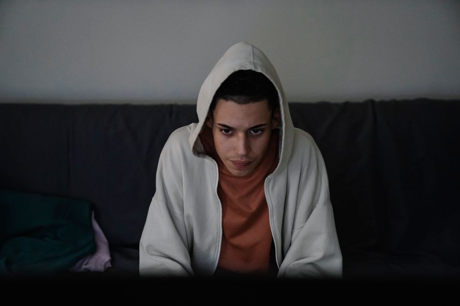 Concentrated young ethnic male in hood looking forward while sitting on sofa in living room