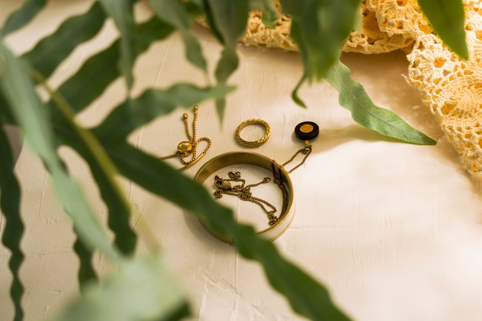 Gold jewelry pieces including bracelets and rings resting on a white textured surface with green leaves and crochet fabric.