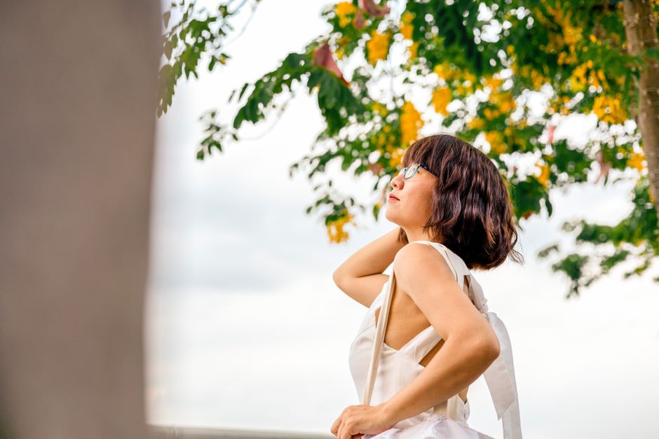 A woman gazes upwards outdoors, with a backdrop of trees and yellow flowers.