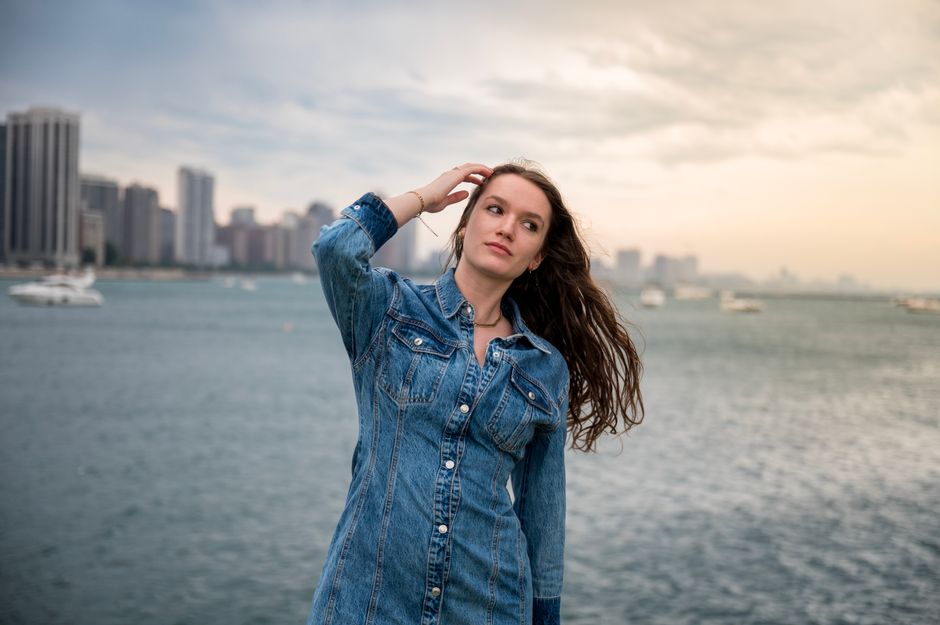 A young woman in a denim jacket poses by a river with a city skyline in the background.