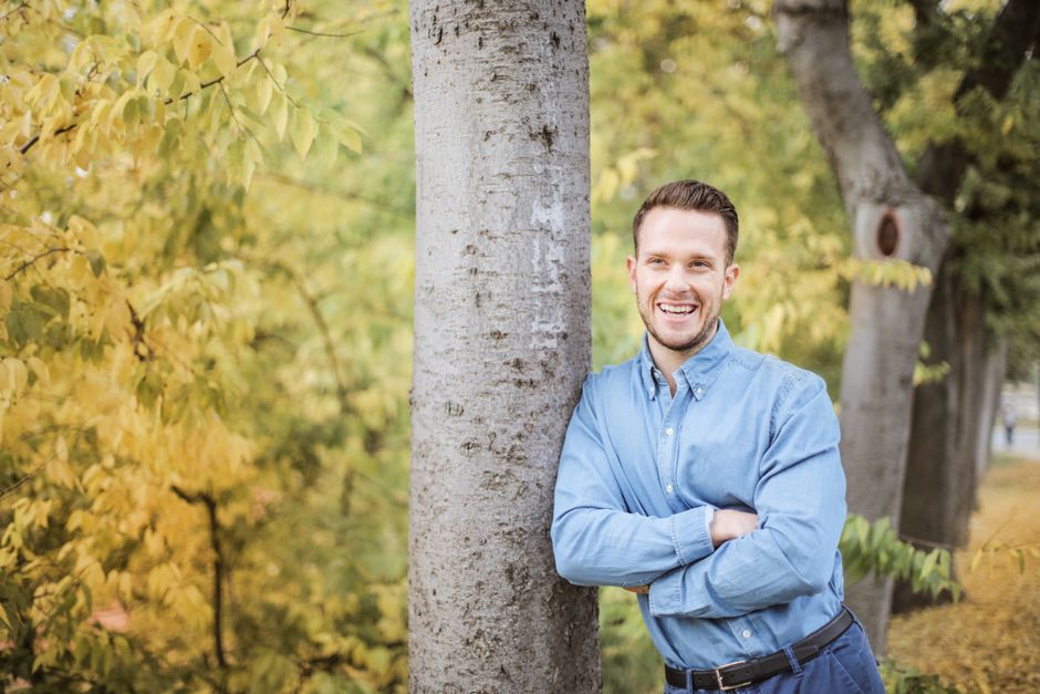 A cheerful man leans against a tree, enjoying the vibrant fall foliage outdoors.
