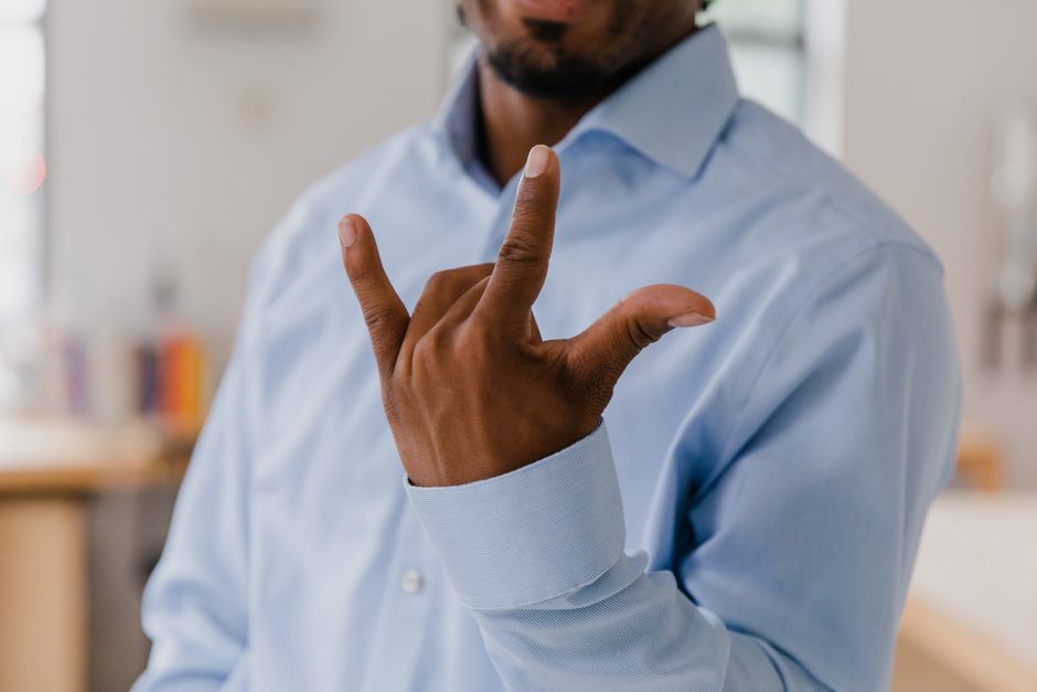 Dark-skinned adult male in a blue shirt making a hand gesture indoors, with focus on hand.