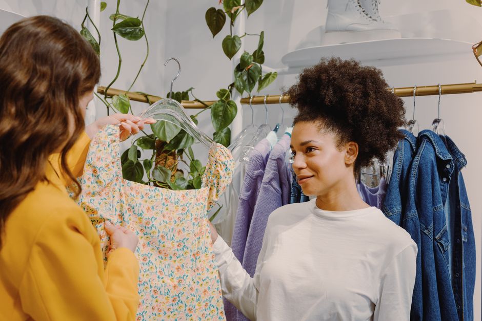 Two women enjoy shopping indoors, exploring a diverse clothing rack with floral prints and denim options.