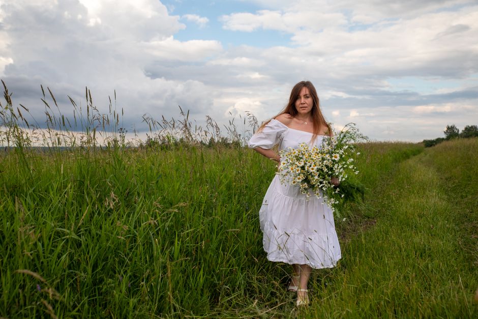 Woman in white dress holding daisies in a summer meadow under cloudy sky.