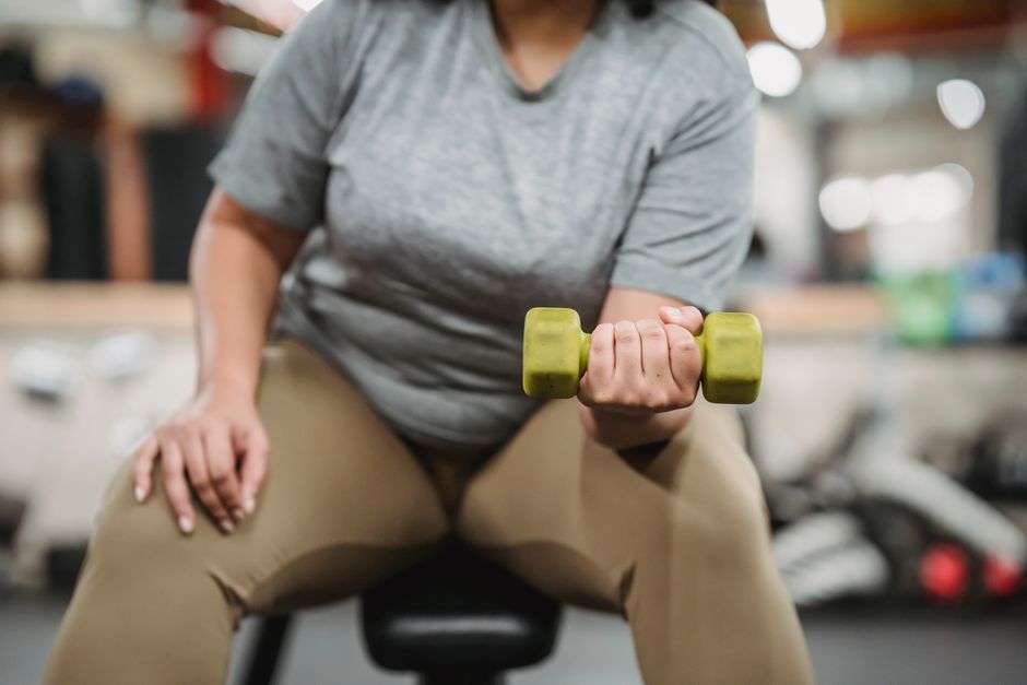 Crop unrecognizable curvy female in sportswear sitting on bench and doing dumbbell curls in gym