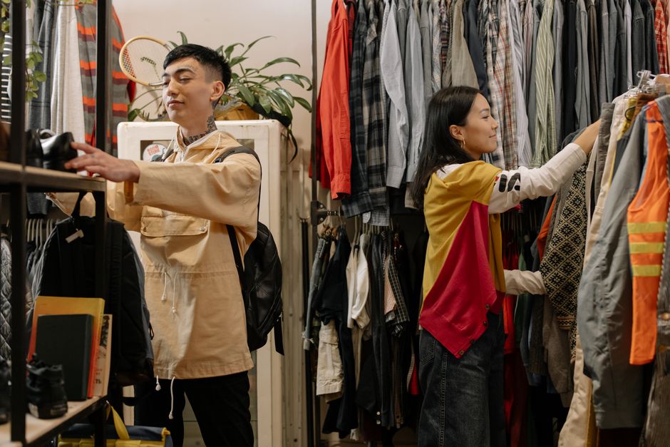 Two young adults selecting clothes in a crowded vintage thrift store.