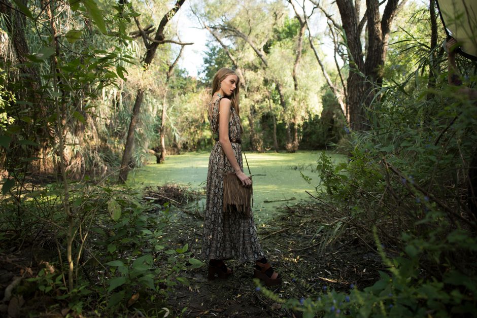 A woman stands elegantly in a forest wearing a patterned dress and fringed bag, captured in a serene natural setting.
