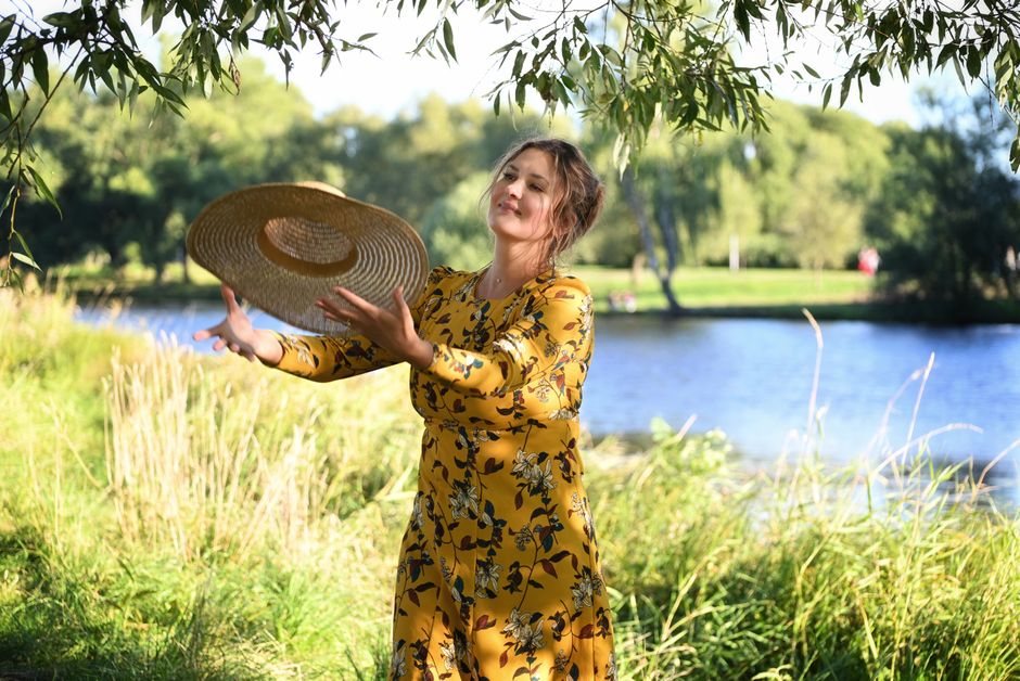 A woman in a yellow floral dress enjoys the outdoors by a river, holding a sun hat.