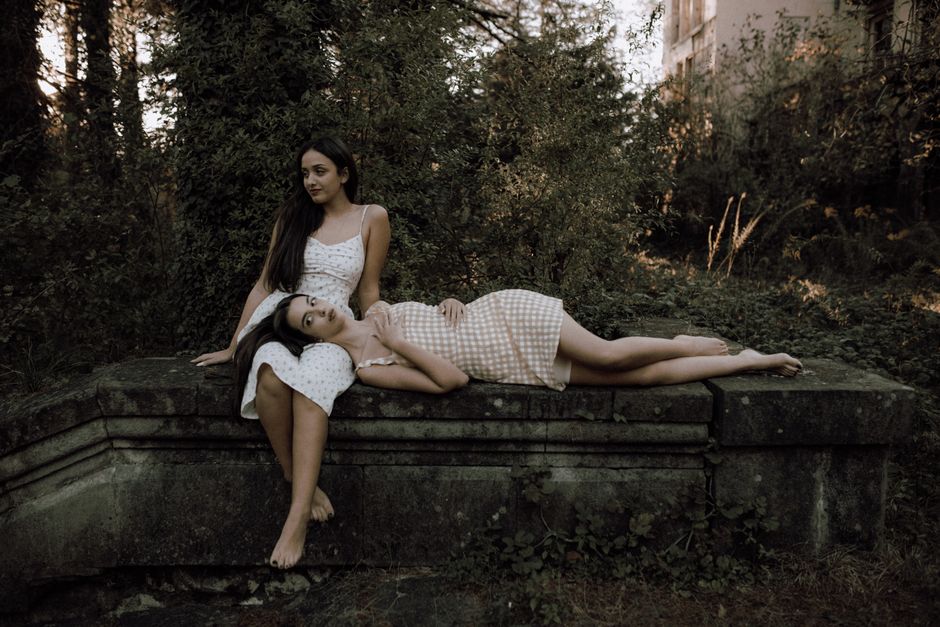 Two women in elegant dresses pose gracefully on a stone bench surrounded by lush greenery.
