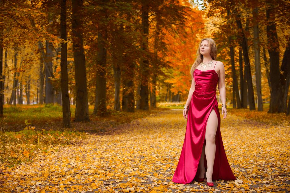 A graceful woman in a red dress stands amidst a vibrant fall forest, surrounded by golden leaves.