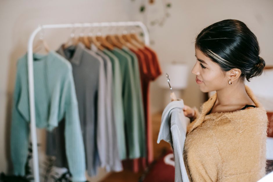 Side view of anonymous ethnic woman in trendy outfit choosing clothes in light bedroom at home with blue blouse on hanger in hands