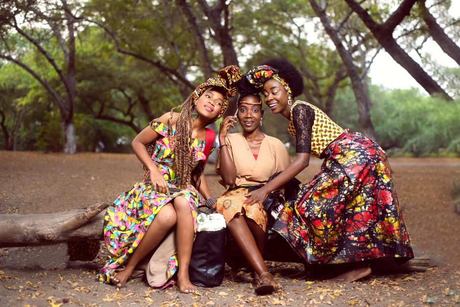 Three women in vibrant traditional attire smiling and posing in a rural outdoor setting.