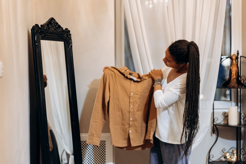 A woman examines a brown shirt in front of a mirror while shopping online at home.