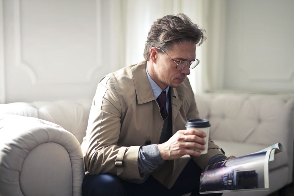 Serious male entrepreneur wearing formal jacket and suit sitting with hot drink to go on comfortable couch in modern flat and reading magazine