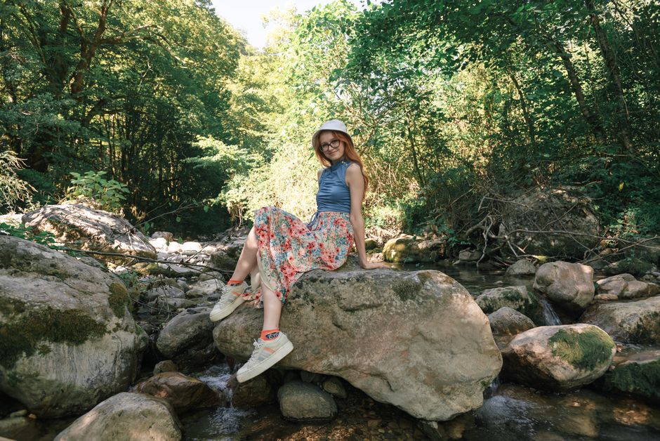 A young woman in a summer dress sits on a rock in a lush green outdoor environment.