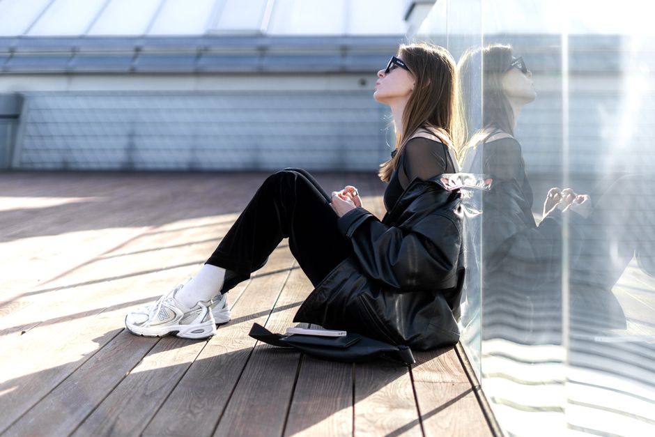 Stylish woman in black outfit sitting against a modern glass wall, reflecting sunlight.