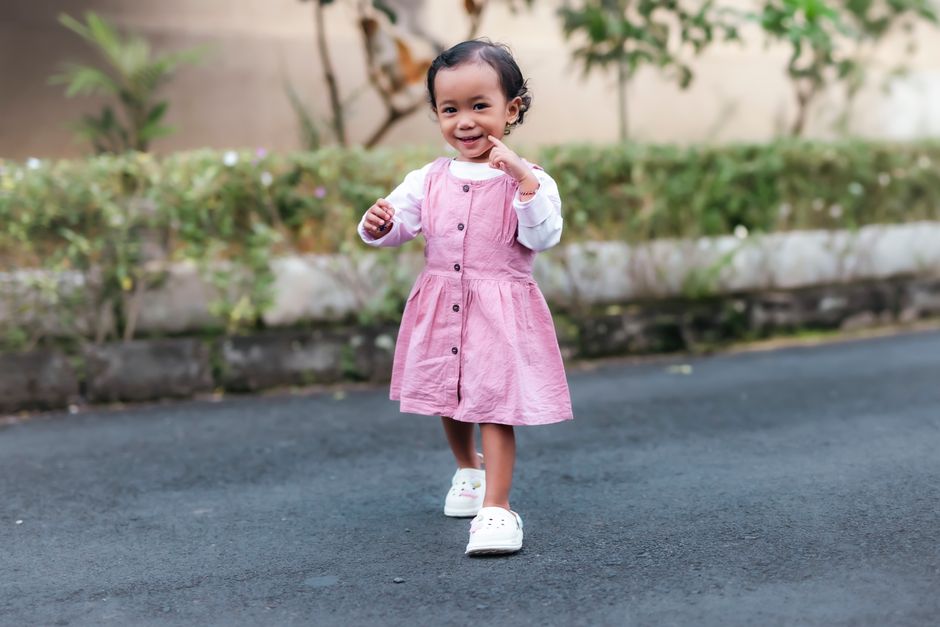 Young child in a pink dress joyfully walking outdoors with a smile.