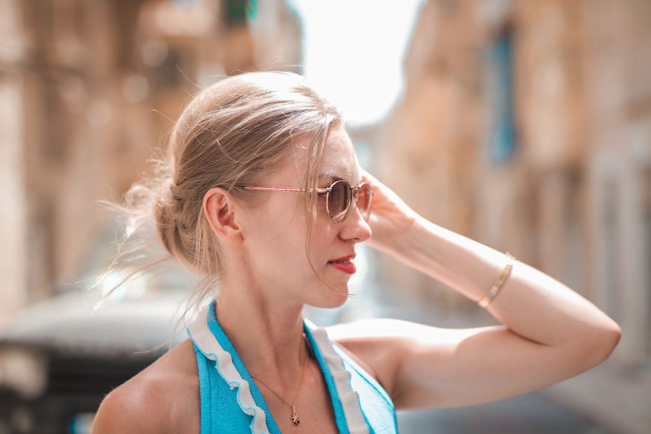 Elegant woman in sunglasses and blue dress stands on a sunlit street in Mgarr, Malta.