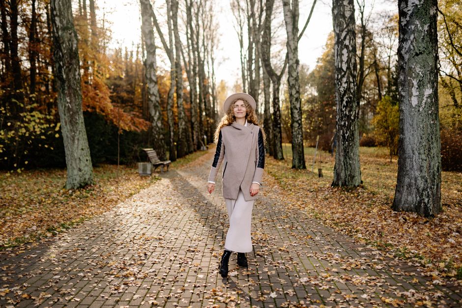 Woman in stylish coat and hat walks along a leaf-covered path in a sunny autumn park.