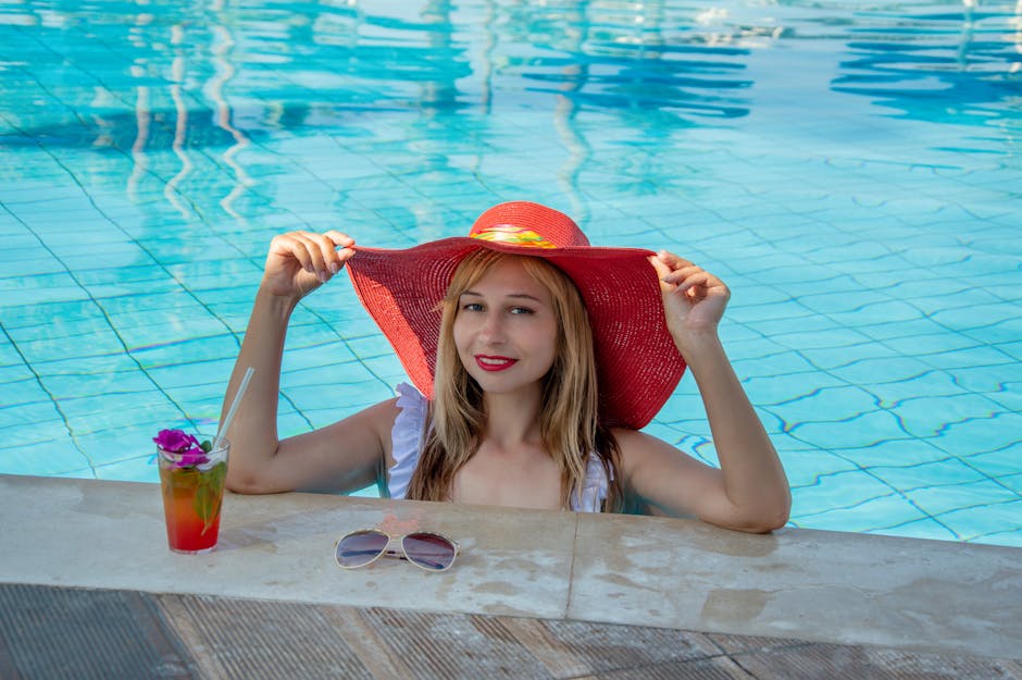 Cheerful woman with red hat enjoys summer by poolside with cocktail and sunglasses.