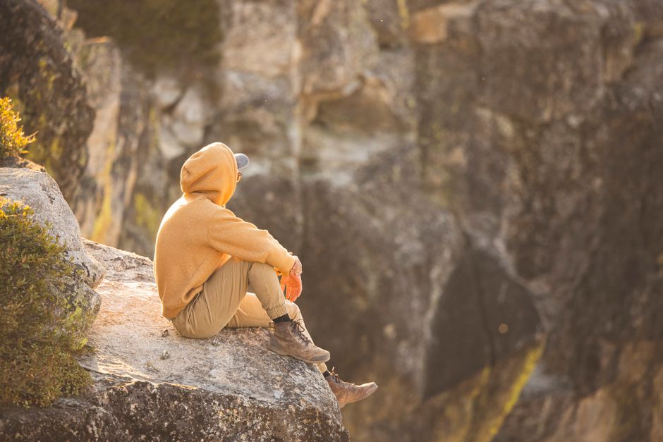 Man sitting on rocky cliff, wearing hoodie and cap, enjoying a peaceful sunset view.